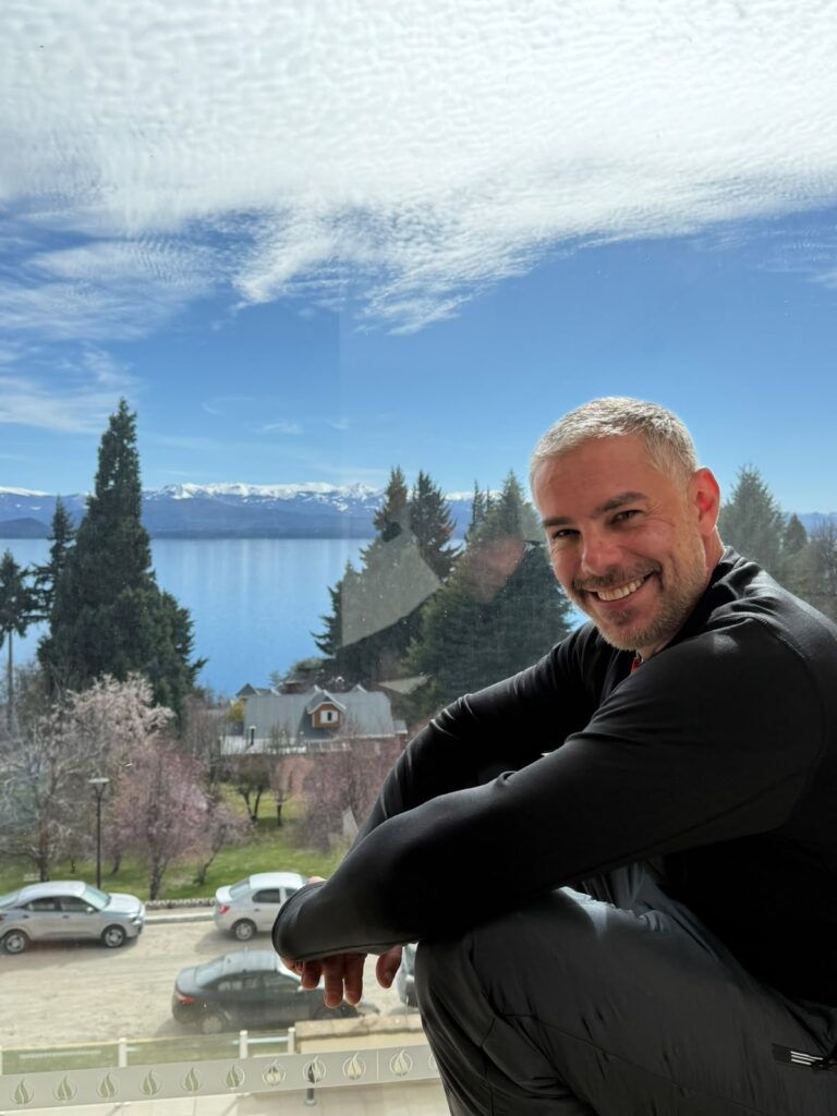 Carlos Eduardo, o Kaká, da KS Turismo, sorrindo durante uma viagem, com lago e montanhas ao fundo.